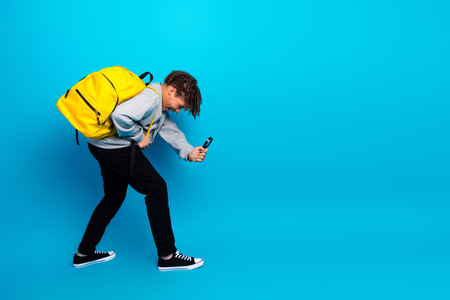 Dynamic young male student in casual style with vibrant yellow backpack and smartphone on a blue backgroundの写真素材
