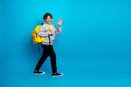 Smiling young boy with yellow backpack waves against a bright blue background, showcasing school and childhood themesの写真素材