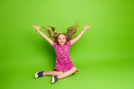 Cheerful young girl in a pink dress smiling and raising arms excitedly against a vibrant green backgroundの写真素材