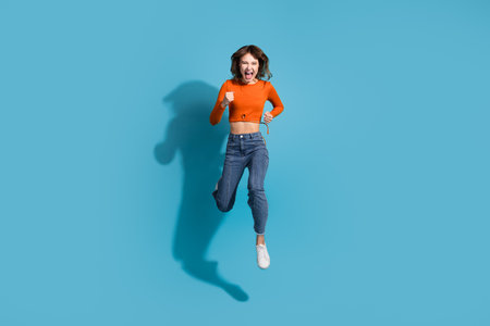 Joyful young woman in stylish orange top and jeans leaping passionately in front of a vibrant blue...