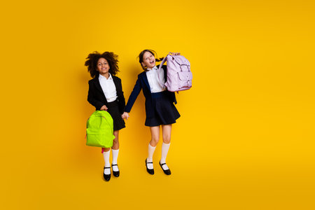 Smiling classmates holding backpacks, jumping on a yellow background, symbolizing friendship, youth, and school diversityの写真素材