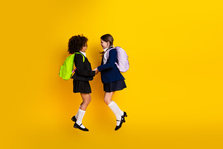 Two cheerful schoolgirls smiling and jumping together on a vibrant yellow background wearing school uniforms and backpacksの写真素材