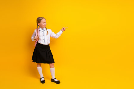 Happy young schoolgirl with blonde braids and backpack pointing with hand, posing on bright yellow backgroundの写真素材