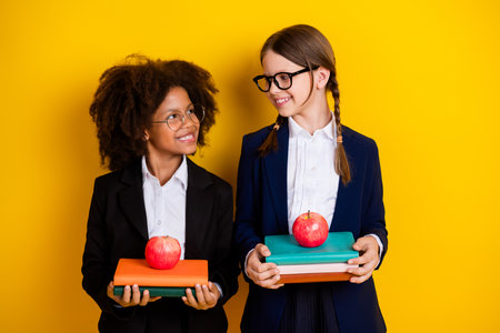 Two cheerful schoolgirls holding books and apples standing in suit uniforms on a yellow background, promoting education.の写真素材