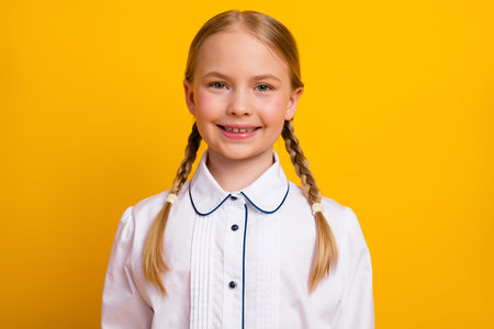 Happy young schoolgirl with blonde braids wearing uniform posing with a bright smile on a yellow backgroundの写真素材