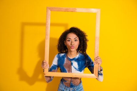 Young woman with curly hair posing playfully holding a wooden frame against a yellow background,...