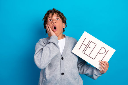 Young schoolboy posing against a bright blue background holding a help sign wearing a grey cardigan...