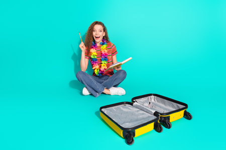 Cheerful young woman with curly hair gestures joyfully while preparing luggage on a vibrant turquoise backgroundの写真素材