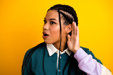Young stylish woman with braids gesturing attentiveness against a vibrant yellow backgroundの写真素材