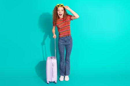 Young woman in striped t-shirt with suitcase on teal backdrop conveying cheerful and trendy travel vibesの写真素材