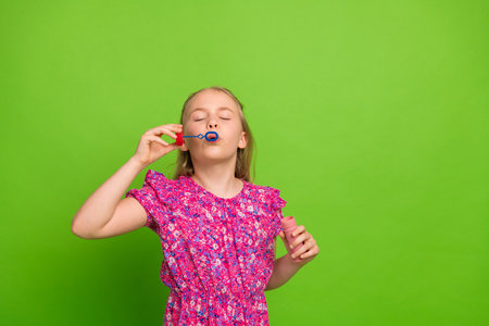 Young girl blowing soap bubbles with a playful expression against vibrant green background promoting...