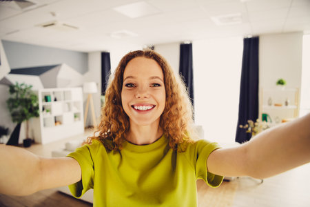 Joyful young woman with curly red hair takes a selfie at home in a stylish living room for a leisure-filled dayの写真素材