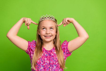 Lovely smiling girl with a crown pointing happily on a vibrant green background during a joyful summer dayの写真素材