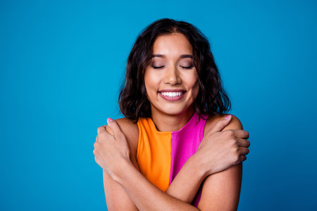 Smiling young woman hugging herself, expressing self love and confidence, vibrant colors on a blue background, joyful momentの写真素材