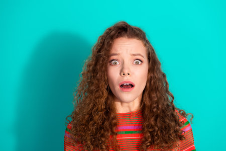 Surprised young woman with curly hair in a striped red shirt against turquoise background expressing amazement and emotionの写真素材