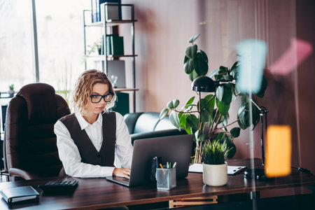 Professional businesswoman working on a laptop in a modern office setting surrounded by stylish decor and indoor plantsの写真素材