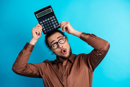 Young businessman in brown shirt holding a calculator over his head choosing solutions on turquoise backgroundの写真素材
