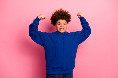 Cute boy in blue hoodie flexing arms against pink backdrop showing confidence and happy expression celebrating funの写真素材