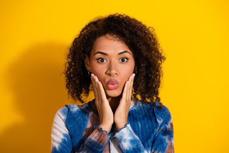 Young woman against yellow background making playful expression, wearing a stylish blue top, showing...