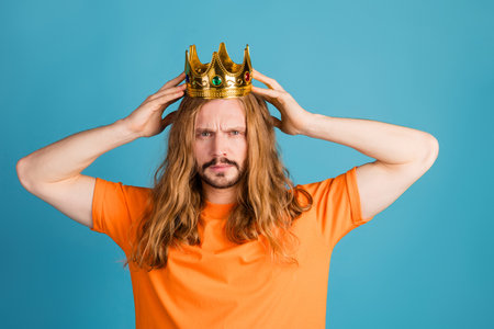 Young man with long blond hair wearing a golden crown poses in a bright orange shirt against a blue backgroundの写真素材