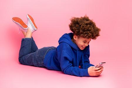 Young boy in blue hoodie using smartphone on pink background showing cheerful expression and modern lifestyleの写真素材