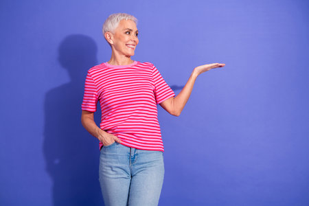 Smiling senior woman in trendy casual wear gesturing with open hand on vibrant purple background