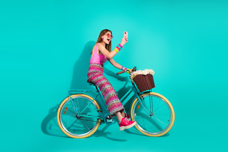 Young woman in trendy casual outfit taking selfie while riding a teal bicycle against a turquoise backgroundの写真素材