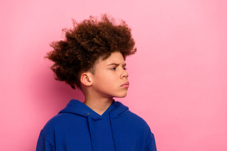 Charming young boy in a blue hoodie standing against a pastel pink background, expressing thoughtful...