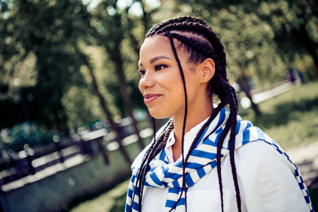 Charming woman with braids enjoying a sunny day outdoors in a city park, wearing casual clothes and...