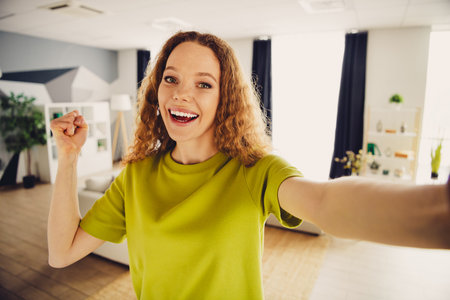 Smiling young woman in a living room confidently posing in a green shirt with a cheerful and uplifting demeanorの写真素材