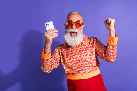 Cheerful senior man in retro fashion attire holding smartphone against vibrant purple background...