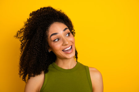Cheerful young woman with curly hair and green top against vibrant yellow background, showcasing casual fashion and styleの写真素材