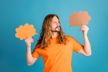 Young man holding speech bubbles in both hands against a blue background, wearing bright clothing...