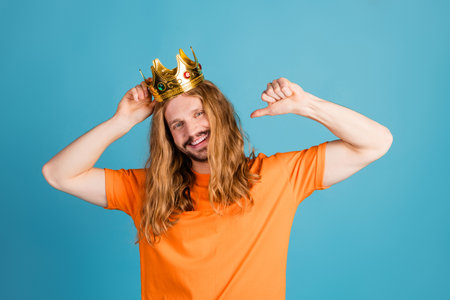 Young man with a golden crown on a blue background gesturing confidently in an orange t-shirtの写真素材