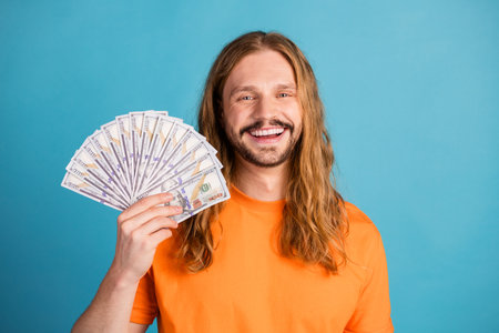 Happy young man holding cash in hand against blue background, demonstrating achievement and positive...