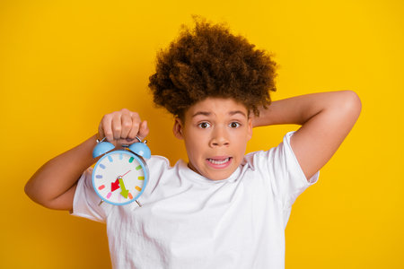 Surprised young boy holding a retro clock against a yellow background, expressing urgency and shock...