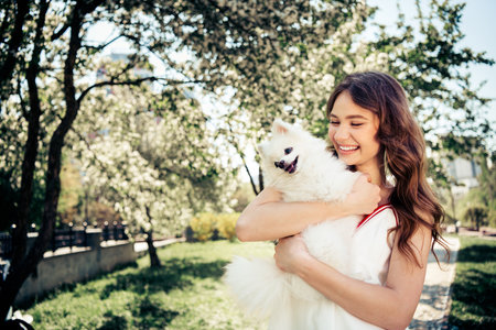 Charming young woman enjoying a sunny day outdoors while holding an adorable white dog in a blooming park.の写真素材