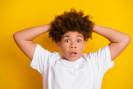 Surprised boy with curly hair posing against a yellow background wearing a casual white t-shirt, expressing amazementの写真素材