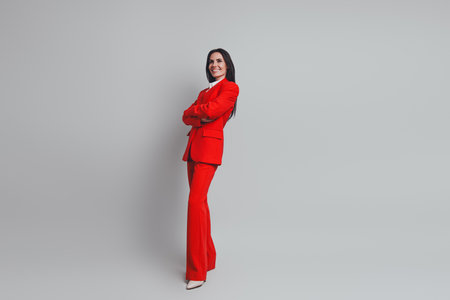 Professional businesswoman in a red suit standing confidently with arms crossed against a grey backgroundの写真素材