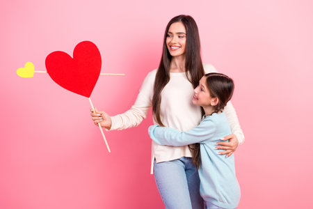 Smiling mother bonding with young daughter holding heart display against pink background illustrating joy and loveの写真素材