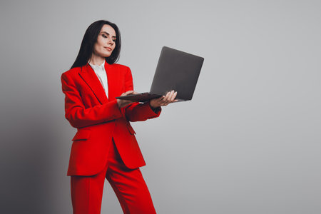 Elegant professional woman in red suit holding a laptop on a neutral grey background, showcasing confidenceの写真素材