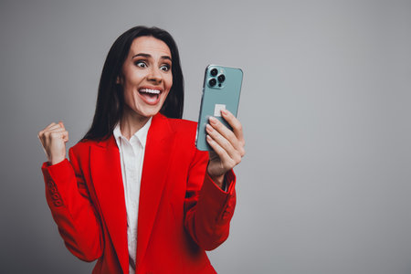 Professional businesswoman celebrating success while checking phone in red suit against grey backgroundの写真素材