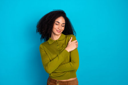 Charming young woman in casual green shirt smiling with arms wrapped in a light blue background for...