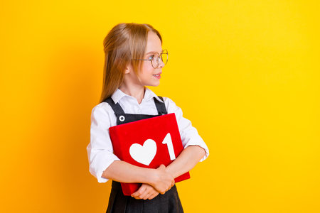 Smiling schoolgirl in uniform holding a red book with a heart symbol against a vibrant yellow...