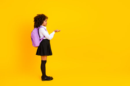 Side profile of young schoolgirl in uniform with backpack open hand gesture bright yellow background education conceptの写真素材
