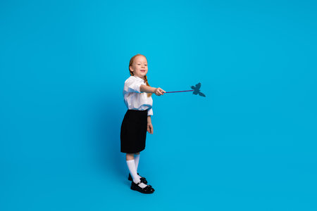 Adorable young schoolgirl with red pigtails poses confidently wearing a charming uniform on a bright blue backgroundの写真素材