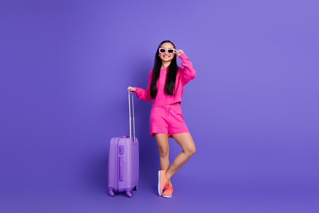 Stylish young woman in vibrant attire with purple suitcase against a bright violet background, embracing trendy fashion.の写真素材