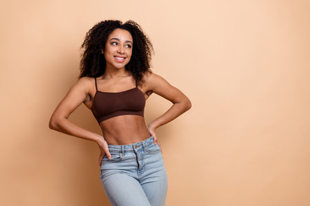 Confident young woman posing on a beige background wearing casual jeans and a brown top, showcasing style and beautyの写真素材