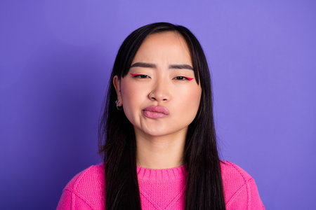 Young multiethnic woman in pink sweater against purple background displaying pensive and comical expression folding lipsの写真素材