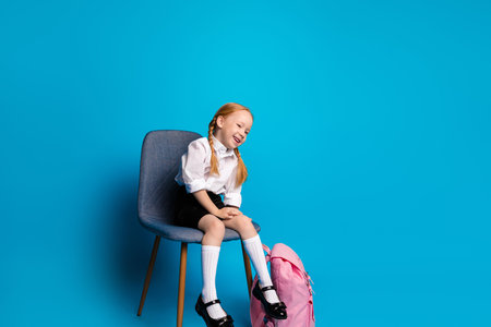 Cheerful schoolgirl sitting on chair with pink backpack on blue background showcasing education and childhood charmの写真素材